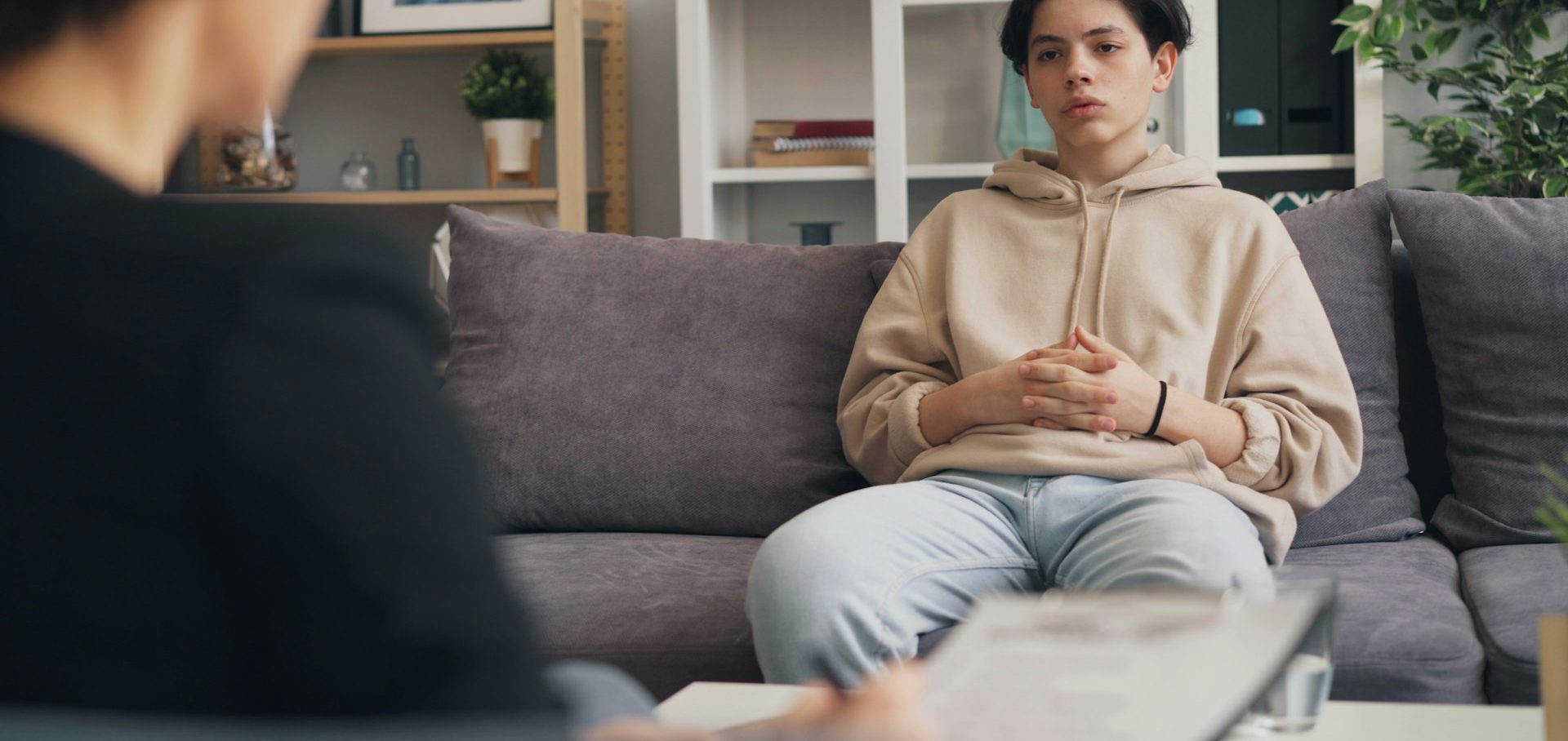 A female with short brown hair is sat on a grey sofa facing someone. Her hands are clasped together in her lap.
