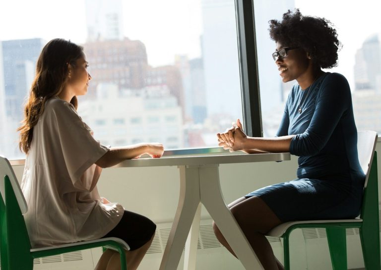 Two females talking at a table. On the left is a white female with long wavy brown hair. On the right is a black female with glasses and an afro. They look like they engaged in a happy conversation.