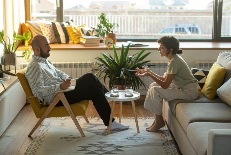 Male therapist speaking with female client. He sits in yellow chair. She sits on grey couch. Looks like an Ikea showroom lots of greys and yellows in this room